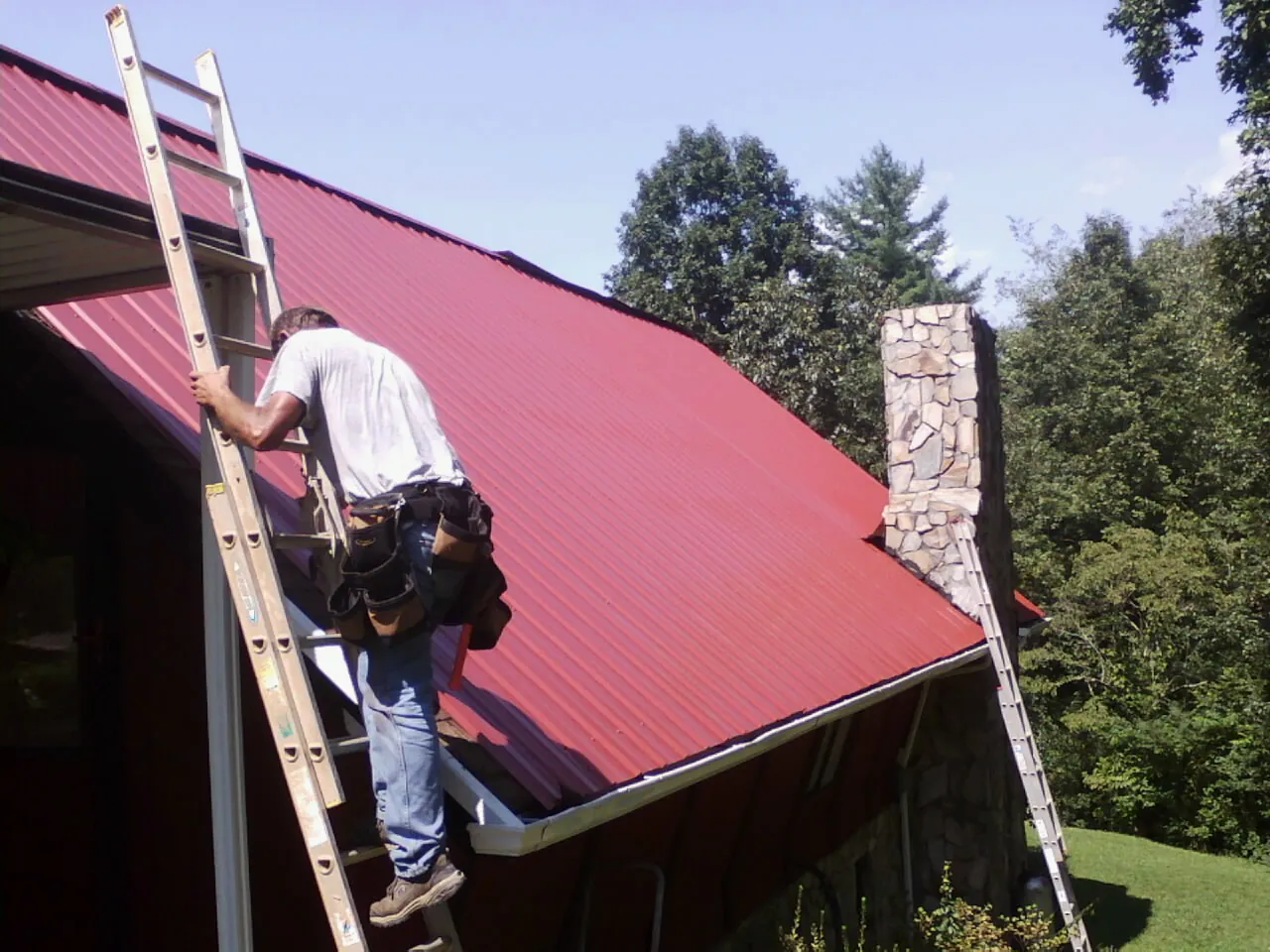 Roofer on a ladder beside a red metal roof and stone chimney — Soffit & Fascia Repair by Roofing Brothers in Harbeson